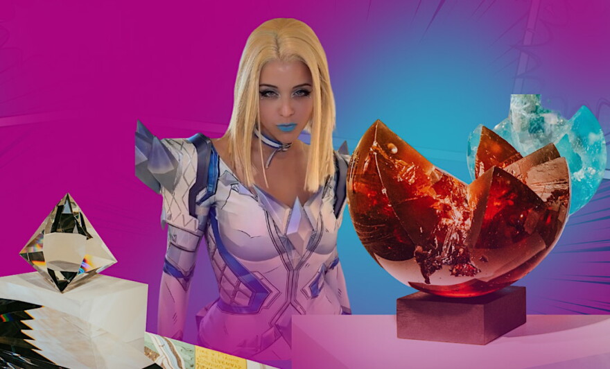 A cosplayer wearing a blonde wig and fake white and blue armor and blue lipstick. There is glass art displayed on both sides of her, on top of a hot pink background.