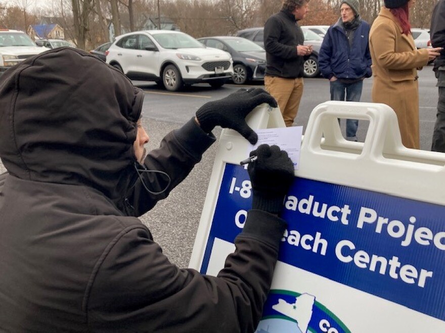 A man in a black winter coat kneels down to fill out a comment card against a sandwich board sign.