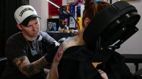 A person lays face down in a massage chair as another person tattoos their back