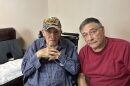 Two men sit side-by-side. One elderly man on the left is in a wheelchair and wears a World War II veterans hat.
