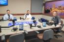 Three men sit at a table in a radio talk studio: a man at left has short dark hair and is wearing a light blue button-down shirt; a man at center has short dark hair and is wearing a white button-down shirt; a man at right has short dark hair and is wearing a grey button-down shirt and jeans.