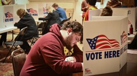 People sit at long tables, bent over while they write. Scattered around them are cardboard dividers that are printed with a waving American flag and read "Vote Here."