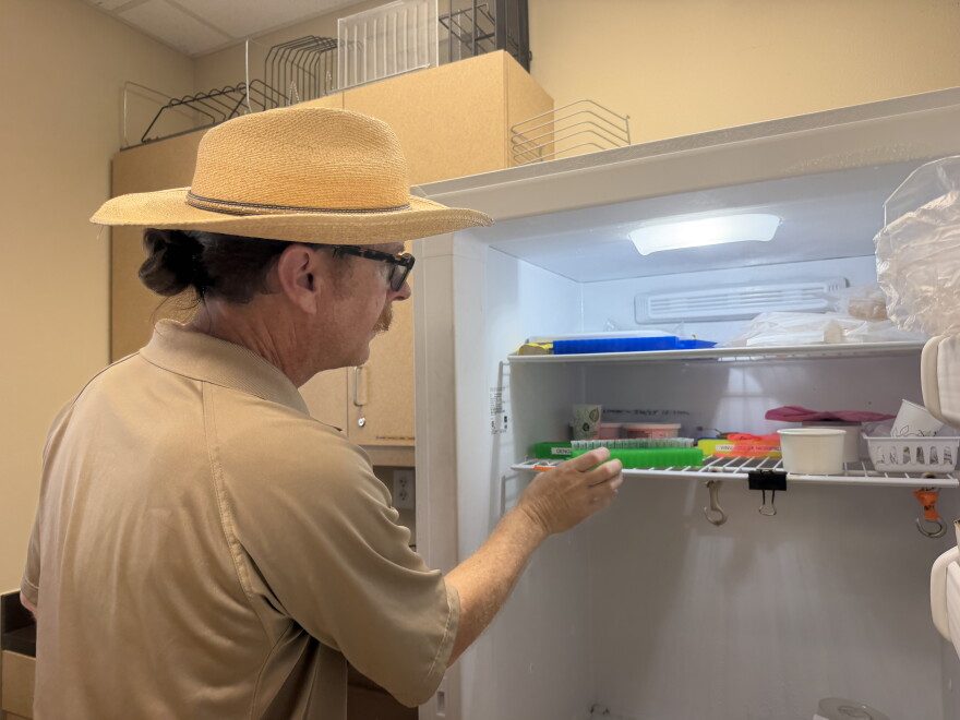 A man in a tan shirt reaching inside a white freezer, grabbing a green tray holding clear test tubes.