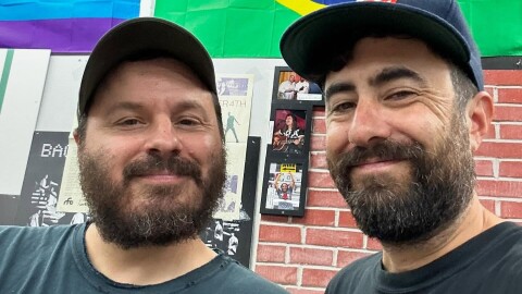 Two white bearded men pose smiling wearing baseball caps with a brick wall in the background adorned with a Brazilian flag on the right corner and Pride flag in the left. There's also some framed music memorabilia on the wall.