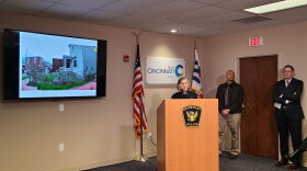 woman stands at a podium. to the viewers left is a TV screen showing body camera still video. It shows a house in a neighborhood in Covington.