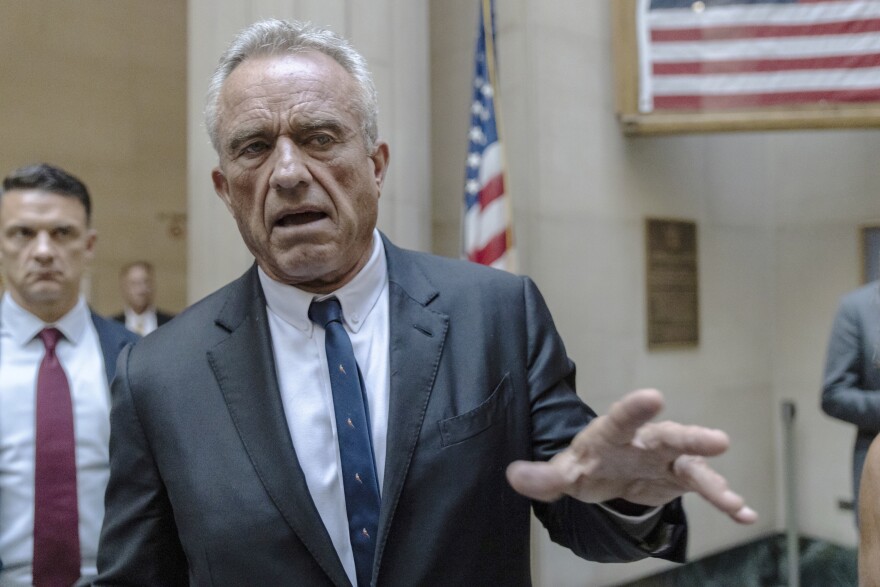 A man in a suit gestures with his left hand in front of a few American flags.