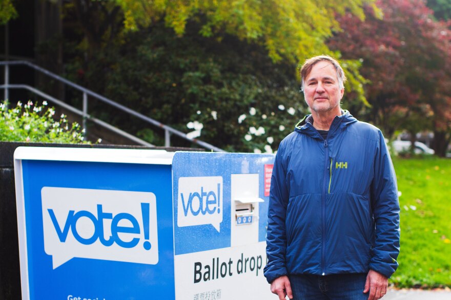 An older man stands next to a blue and white ballot drop box with his hair graying above either ear while wearing a blue jacket, jeans and white tennis shoes.