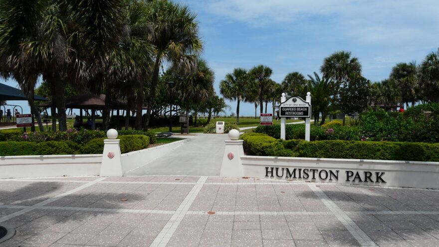 Entrance to Humiston Park in Vero Beach, Florida, showing a paved plaza leading to a walkway lined with palm trees and shrubs, with the park's name carved into a stone wall on the right and a sign for guarded beach access and restrooms in the background.