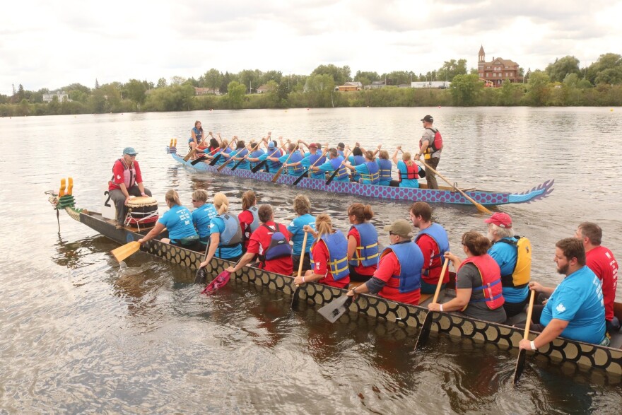 Two long boats containing over a dozen people with paddles are lined up side to side in the water with a green shoreline in the background