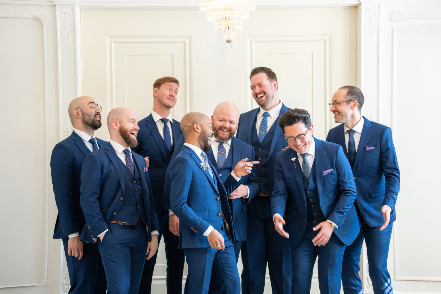 The eight-person Cantus vocal group standing, laughing, wearing matching blue suits in front of a white wall