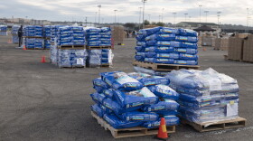 Stacks of blue bags of pet food are arranged on pallets in a parking lot. The sky is overcast and there are tall stadium lights in the background.