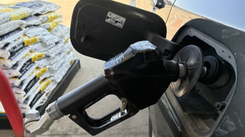 A black gas pump nozzle is inserted into a gray SUV with a pallet of bags of ice melting compound visible in the background. 