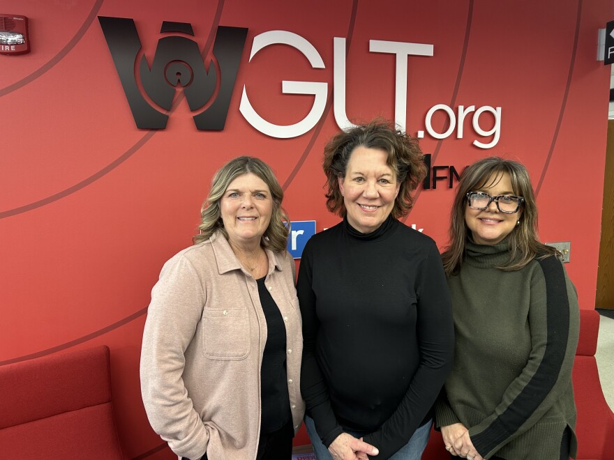 Three women stand shoulder to shoulder smiling at the camera. They stand in front of a large red wall. 