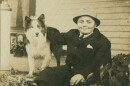 A monochromatic photograph of a young Herman B Wells wearing a dark suit jacket and a brimmed hat, seated outdoors while resting his arm on his standing, long-haired collie dog.