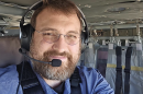 A man in glasses and a slightly graying beard smiles to the camera. He's wearing a headset and looks to be strapped in to a seat in a helicopter or similar vehicle.