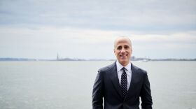A smiling man with short grey hair wearing a black suit, white collared shirt, and black tie with white polka dots stands in front of a river with the Statue of Liberty in the background