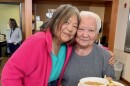 Two women stand close together, one holding a plate with bread and a bowl of soup.