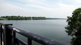 A serene view of a calm Scioto River from a bridge, with a railing in the foreground. The river is surrounded by lush green trees, and the sky is partly cloudy.
