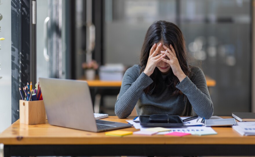 A woman with long dark hair sits with her head in her hands at a desk covered in devices and papers.