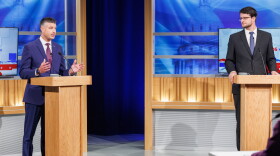 Two men in suits stand at podiums in a TV studio
