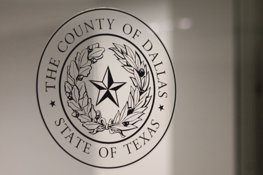 A transparent version of the Dallas County seal on a glass door. The seal includes the words "The County of Dallas - State of Texas" as well as a star in the center of two curved branches of leaves.