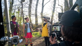 two men and a woman stand in the forest while a TV camera crew films. One of the men is in a yellow and black bird costume