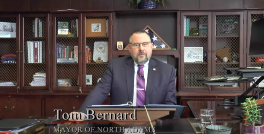 A white man with a beard and glasses sits behind a desk in a suit in front of a wood and glass bookshelf.