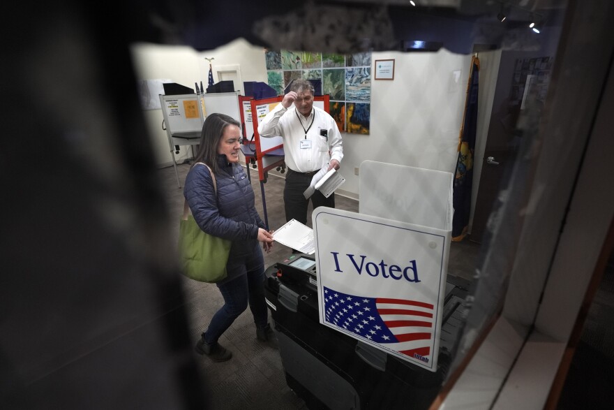 Viewed through glass, a person places a ballot in a ballot-counting machine marked with an American flag and the words "I voted" while another person looks on