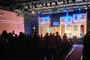 A crowd of people at a theater standing up clapping for a stage with three people and a fake street behind them.