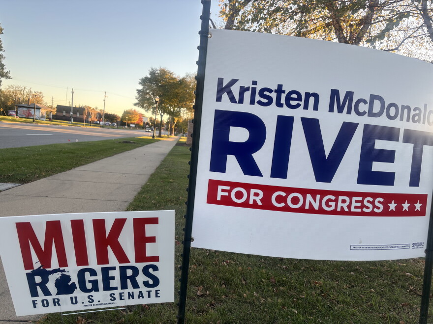 Two campaign yard signs stand on a grassy lawn next to a sidewalk and empty street. On the right, a large white sign reads "Kristen McDonald RIVET FOR CONGRESS" in bold blue and red lettering with three stars. To the lower left, a smaller sign reads "MIKE ROGERS FOR U.S. SENATE" with a small blue silhouette of Michigan built into the text. Trees and buildings are visible in the background under a clear sky.