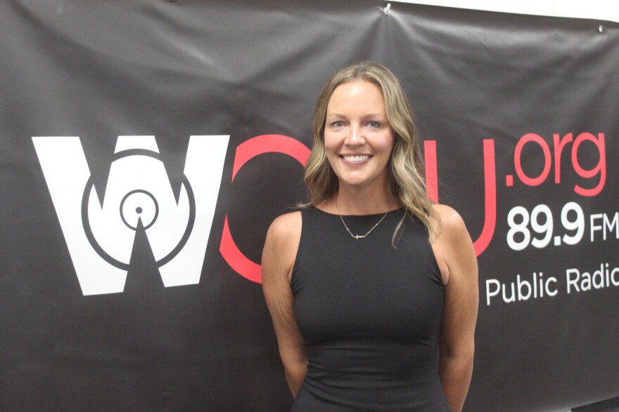  Nicole stands in front of a black poster that says WCBU.org public radio. She wears a black dress, and has blonde hair. 