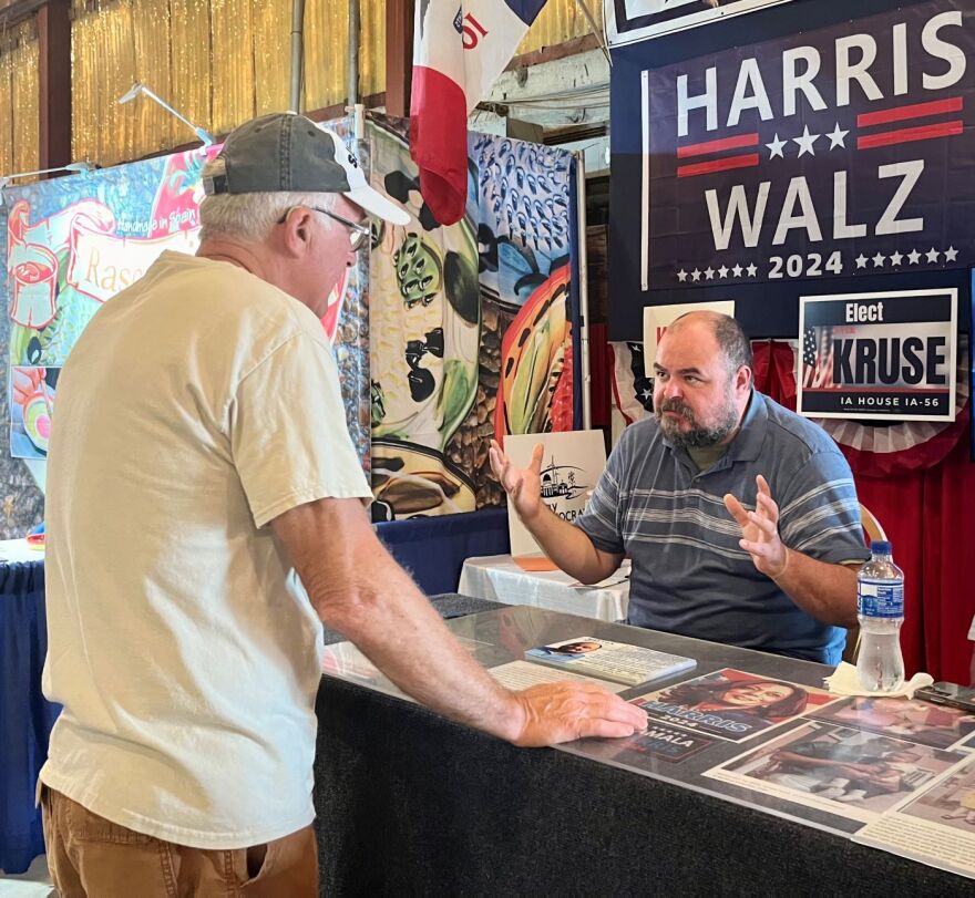 Two men are speaking at a booth for the Clay County Democrats at a county fair. A man is wearing a white t-shirt and has his back to the camera. A man with balding hair and brown beard is to the right. He has his hands up and is engaged in lively conversation.