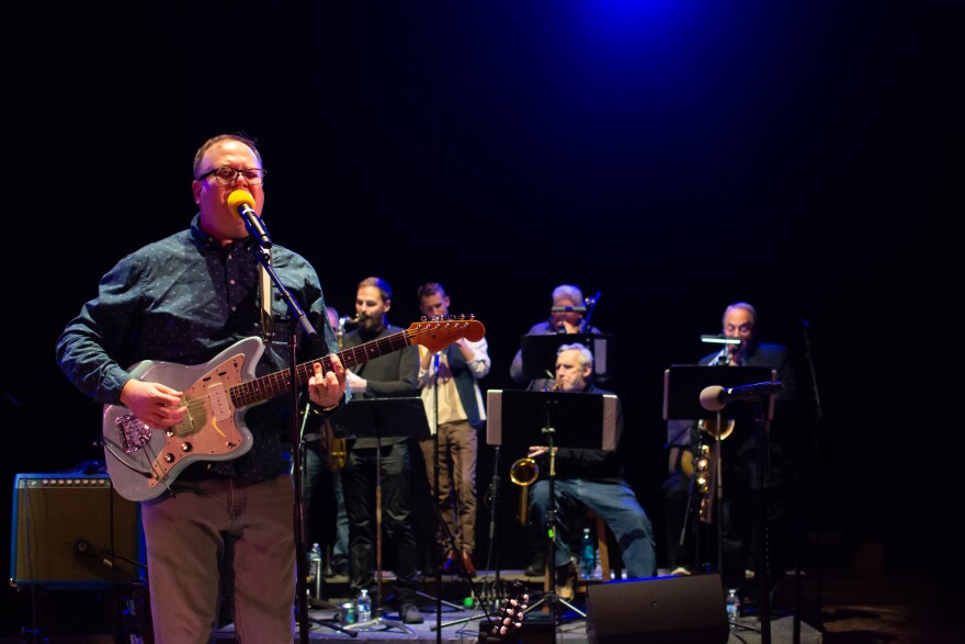 Local musician Rich Reuter plays a guitar and sings in the foreground with a horn section blurred in the background
