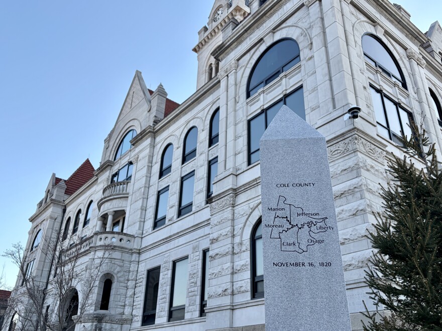 The white brick, gothic architecture Cole County courthouse exterior is in the background. A pillar with a map of Cole County is in the foreground. 
