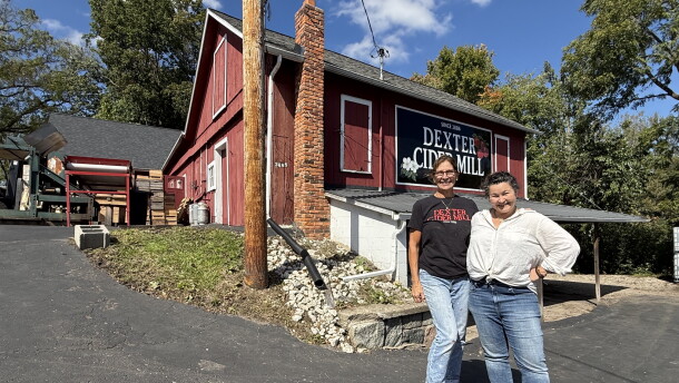 two women stand on a black driveway in front of a red barn that has a "Dexter Cider Mill" sign on it