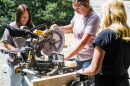 A young woman with a ponytail and clear work goggles uses a saw, with sawdust flecks flying in the air around her, as two other women stand watching. 