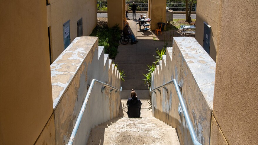 A person with their back to the camera sits midway down an outdoor staircase. Other people can be seen in the shade in the distance