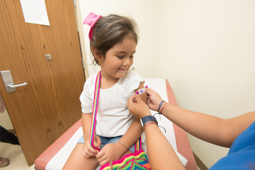 A small child smiles and sits on a chair as a healthcare worker wearing blue gloves places a bandage on the child's arm.