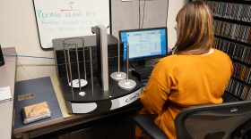 woman in gold dress sits at computer with CD ripping machine to her left, in a CD library