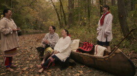 Four individuals smile and laugh while in a forest near a body of water. Two of the individuals are leaning against a boat, while the other two are standing. 
