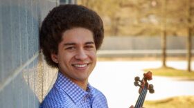 Violist Dillon Scott wearing a blue checked shirt, holds his viola and leans against a building wall outside.