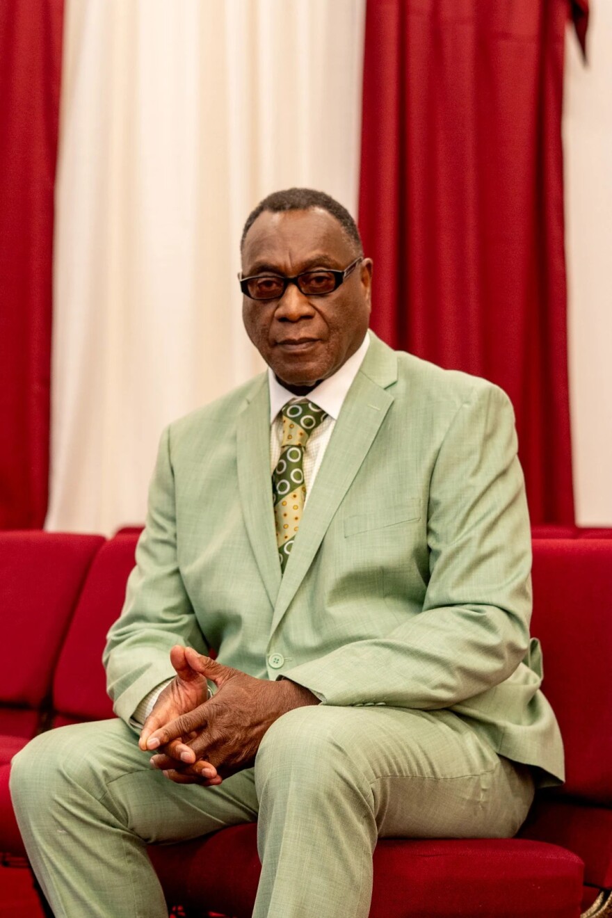 A man in a light green suit sits on a red sofa, hands folded in front of him.