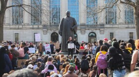 Hundreds of students gather by a large statue and an academic building. They hold signs, including one that reads "D-E-I or DIE" 