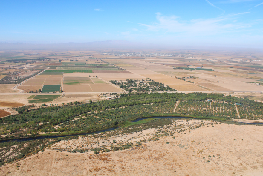 Aerial view of a mostly yellow and brown landscape with a path of green following near where a river flows through. You can visibly see where land plots are sectioned off and more development in the background.