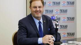 Eddie Gossage, dressed in a suit and sitting in front of a microphone at a press conference.