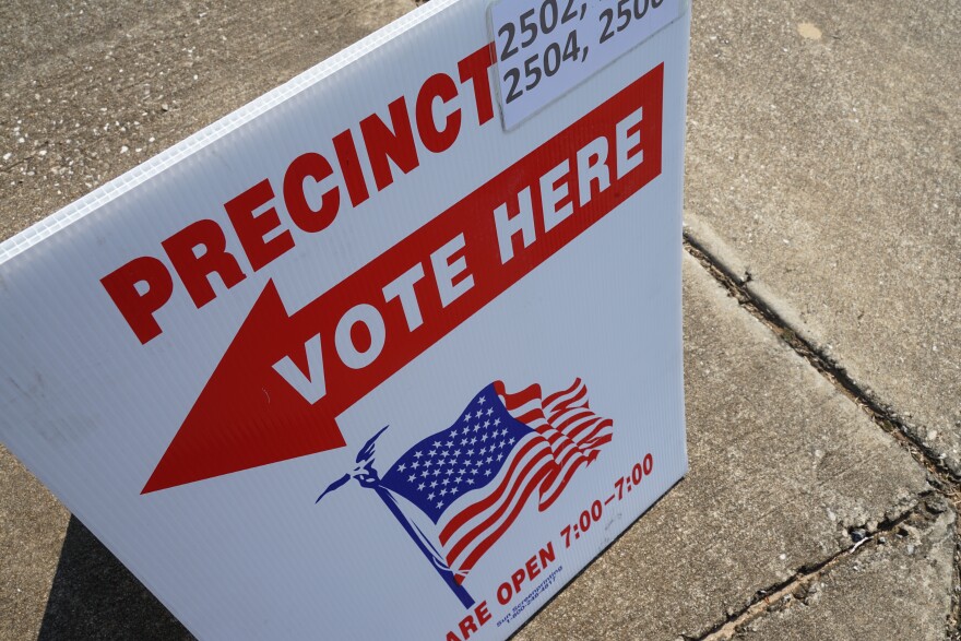 large sign is set up on the sidewalk. The sign reads, "Precinct." Then, an arrow points to the left. The words, "Vote here" are written on the arrow. Below the arrow is an American Flag and below the flag is the words, "We are open 7:00 - 7:00." 