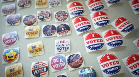 Close up image of about 30 different lapel stickers lying flat on a table. Some read "I Voted Today" with a red, white and blue design. Others read "Be a Voter" in colorful designs.