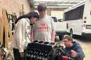 Two students stand over a car engine while a man explains the mechanics.
