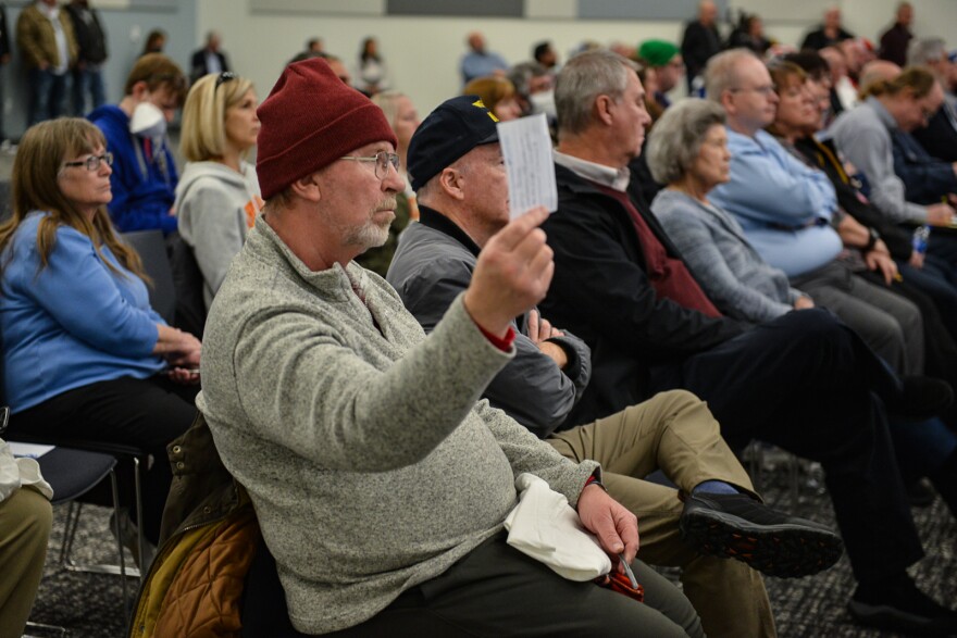 A man sitting in a row of chairs in an audience holds up with his right hand an index card waiting for someone to retrieve it.