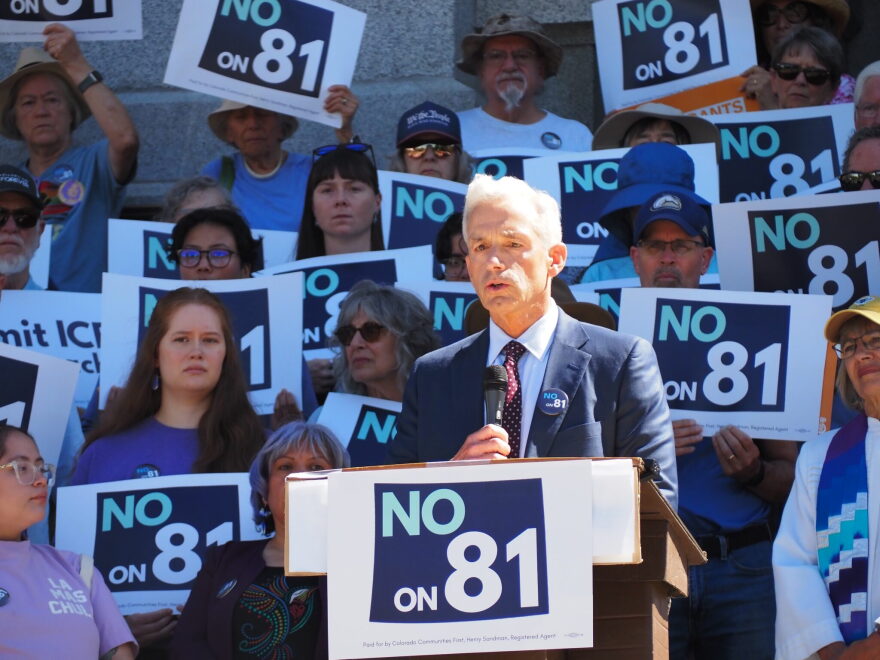 Denver District Attorney John Walsh speaks at a podium surrounded by people holding signs that say "No on 81"